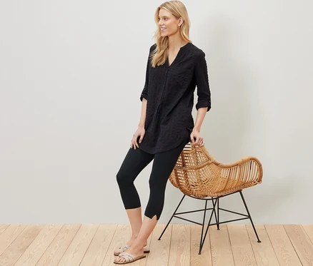 Blonde Frau lehnt an einem Rattansessel und trägt eine schwarze 3/4-Leggings mit Detail.