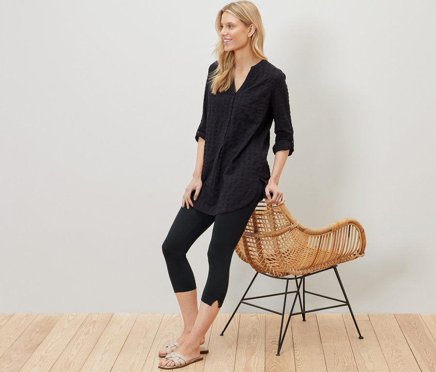 Blonde Frau lehnt an einem Rattansessel und trägt eine schwarze 3/4-Leggings mit Detail.