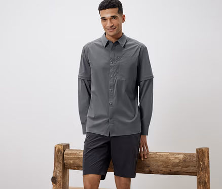 Mann, der sich an eine Holzbank lehnt, trägt ein graues Outdoorhemd und schwarze Shorts.