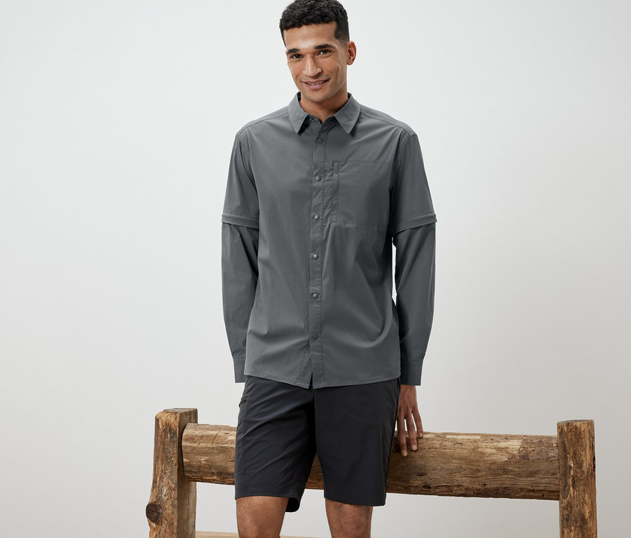 Mann, der sich an eine Holzbank lehnt, trägt ein graues Outdoorhemd und schwarze Shorts.