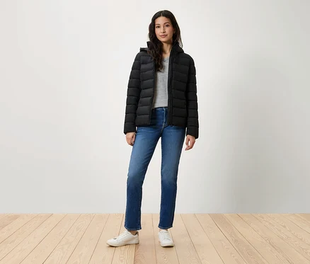 Eine Frau steht und trägt eine schwarze Daunenjacke, blaue Jeans und weiße Turnschuhe.