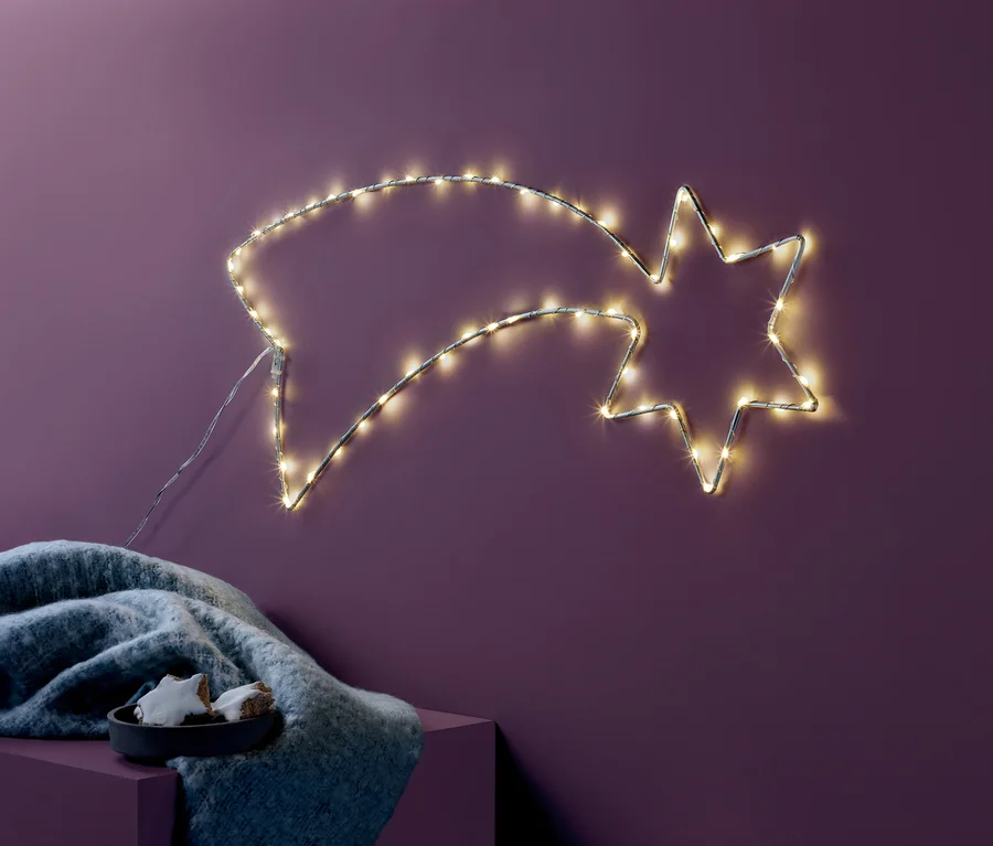 An einer lila Wand hängt eine LED-Sternschnuppe. Darunter liegen auf einem lila Würfel eine graue Decke und eine Schale mit Süßigkeiten.