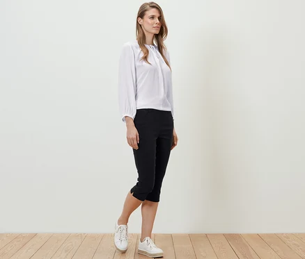 Eine Frau steht auf einem Holzboden und trägt eine weiße Bluse und eine schwarze 3/4-Bengalin-Hose.