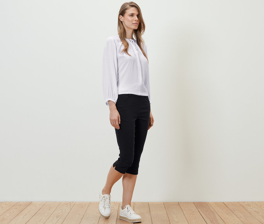 Eine Frau steht auf einem Holzboden und trägt eine weiße Bluse und eine schwarze 3/4-Bengalin-Hose.