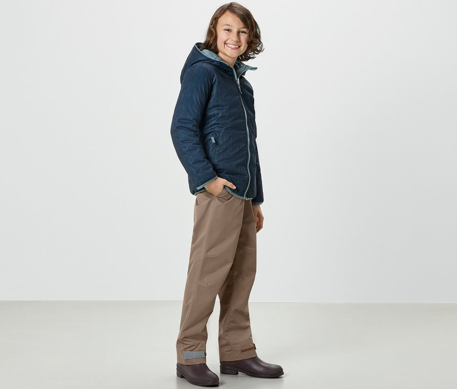 Ein Junge in Jacke und Kinder-Regenhose steht seitlich.