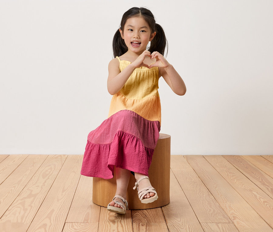 Mädchen sitzt auf einem Hocker und formt mit den Händen ein Herz. Sie trägt ein Kinder-Webkleid.