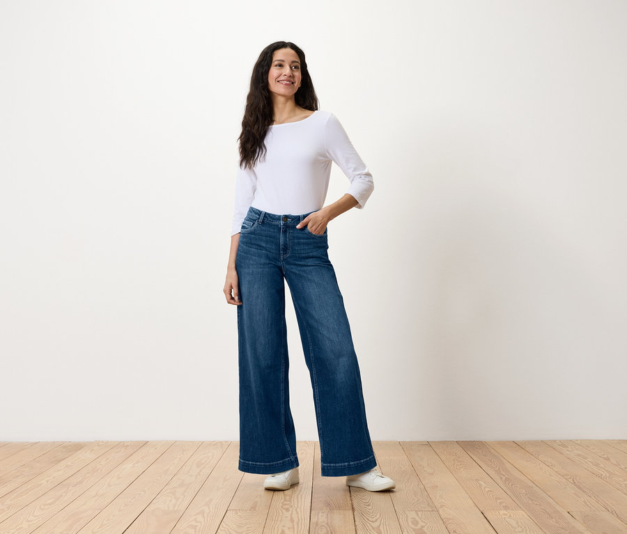 Frau steht und posiert in locker sitzenden blauen Jeans und einem weißen Oberteil.