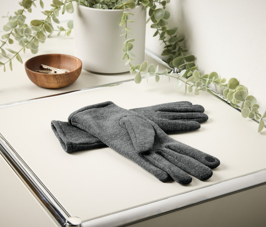 Graue Handschuhe liegen auf einem weißen Tisch. Im Hintergrund befinden sich eine hölzerne Schale mit Schlüsseln und eine Blume in einem weißen Blumentopf.