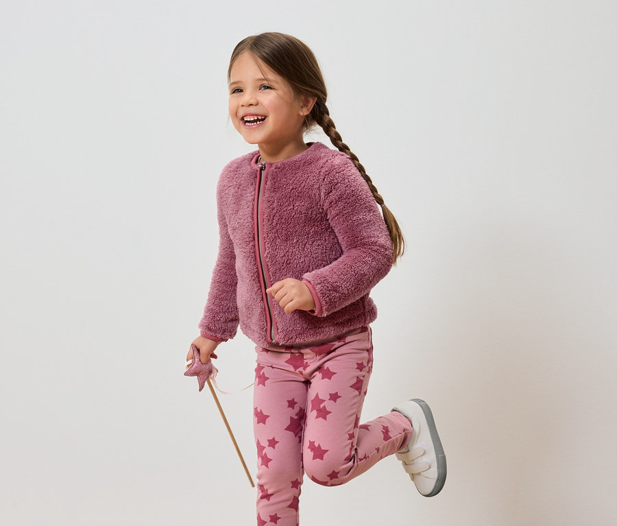 Lächelndes Mädchen mit braunen Haaren in rosa Jacke und Hose mit Sternen hält einen Zauberstab.