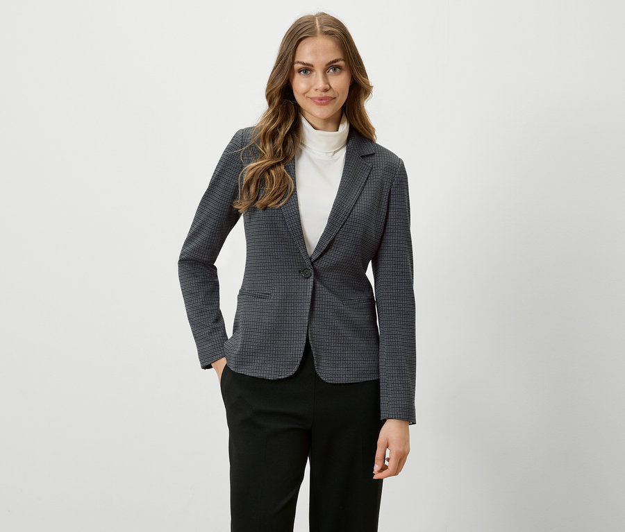 Frau mit braunen Haaren trägt einen karierten Blazer und schwarze Hosen.