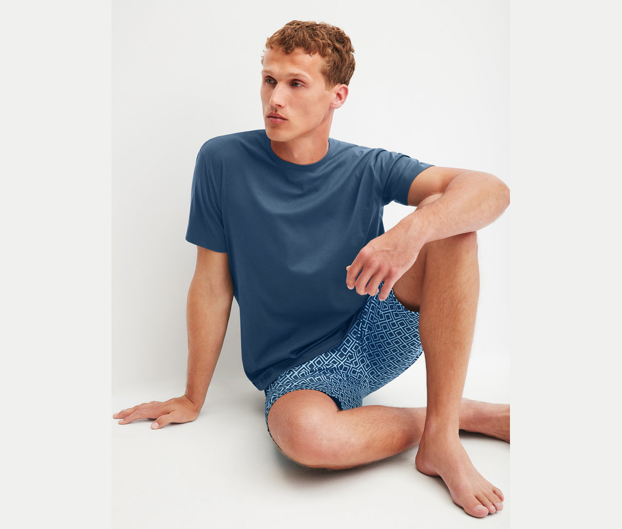 Mann sitzt auf dem Boden und trägt ein blaues T-Shirt und blaue Shorts mit geometrischem Muster.
