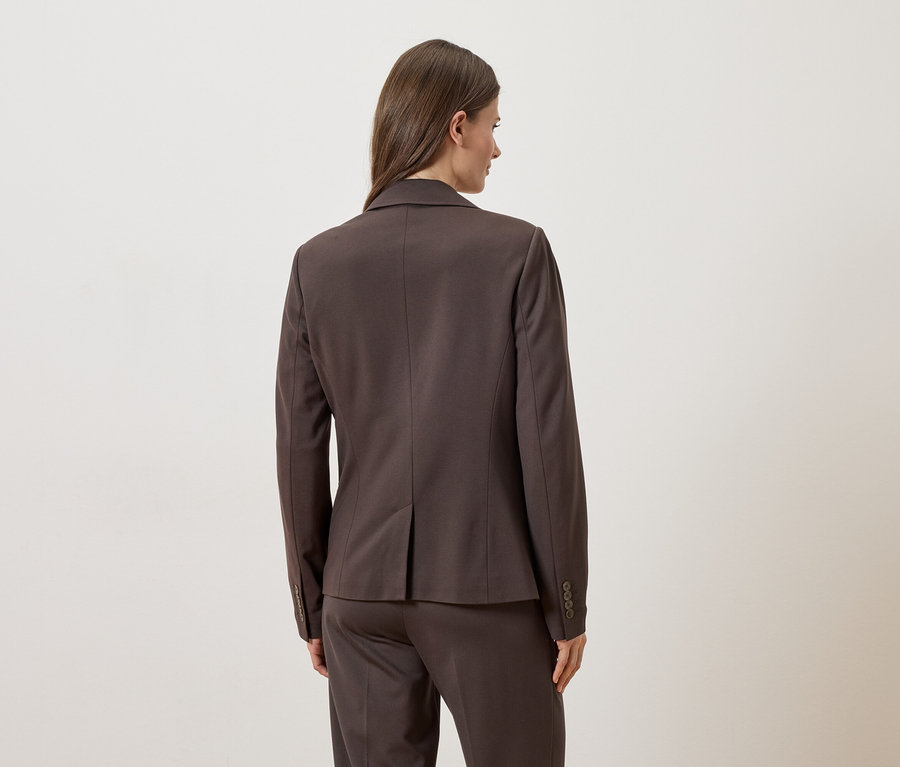 Rückansicht einer Frau mit braunem Blazer und Hose vor einem weißen Hintergrund.