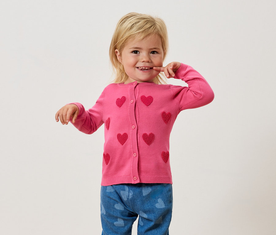 Ein blondes Mädchen trägt eine pinke Strickjacke mit Herzmuster und blaue Jeans mit Herzmuster.