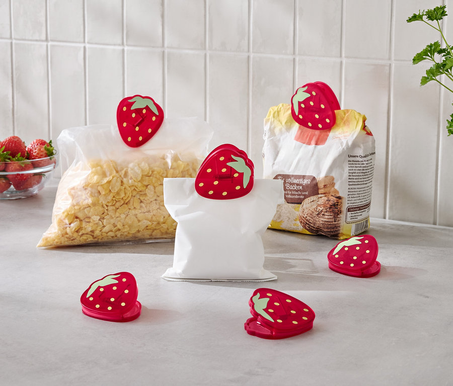 Auf einer Küchentheke stehen eine Schüssel Erdbeeren, eine Tüte Cornflakes, eine Tüte Mehl und eine leere Tüte, die alle mit roten Erdbeer-Clip-Verschlüssen verschlossen sind.