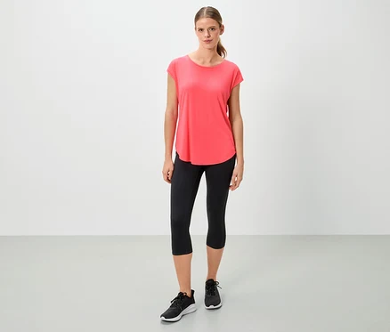 Frau in einem korallfarbenen T-Shirt, schwarzen Leggings und Turnschuhen steht vor einem weißen Hintergrund.