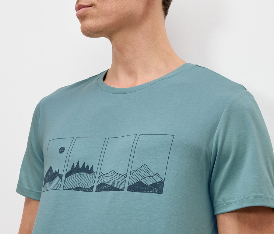Ein Mann trägt ein türkisblaues T-Shirt mit einer Grafik von Bergen und Bäumen in Rechtecken.
