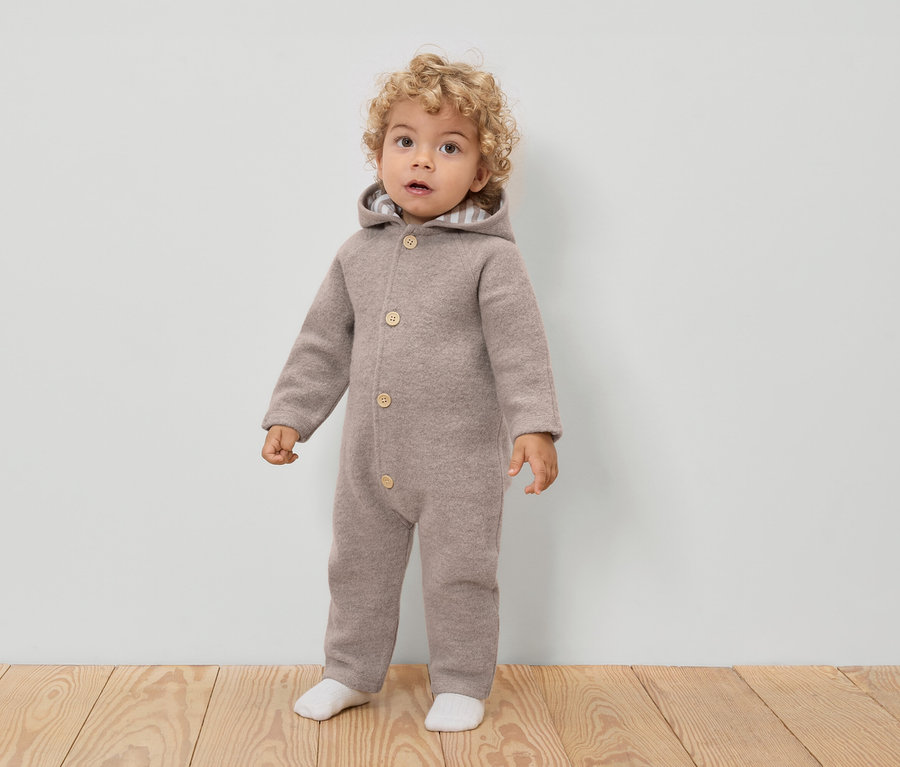 Ein Kleinkind mit lockigen Haaren trägt einen hellbraunen Woll-Overall mit Kapuze und Knöpfen, dazu weiße Socken.