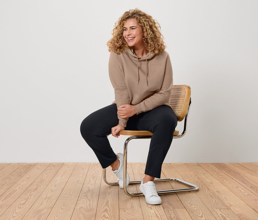 Frau mit lockigem Haar sitzt auf einem Stuhl, trägt einen beigen Hoodie, schwarze Hose und weiße Turnschuhe.