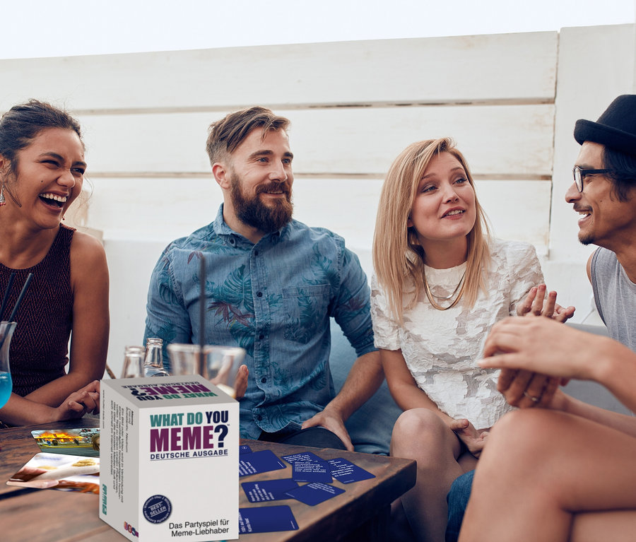 Vier Freunde sitzen an einem Tisch mit dem Spiel »What do you meme«. Karten liegen auf dem Tisch.