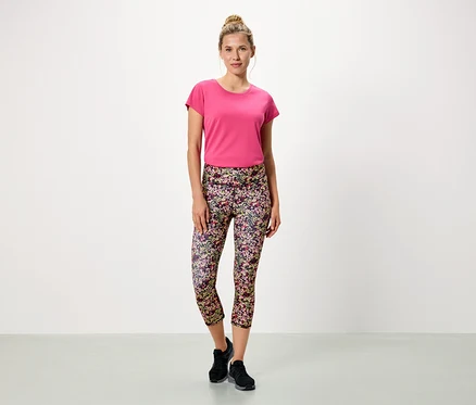 Frau steht vor einem weißen Hintergrund und trägt ein pinkes T-Shirt und blumige Leggings.