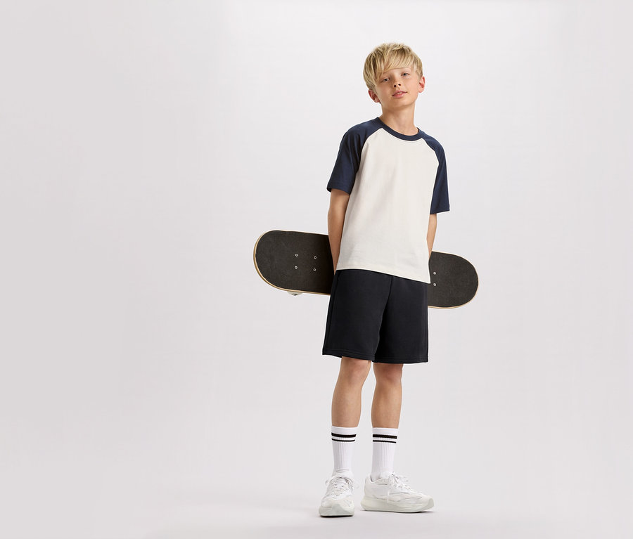 Ein blonder Junge hält ein Skateboard hinter seinem Rücken, trägt ein T-Shirt mit marineblauen Ärmeln, Shorts und Turnschuhe.