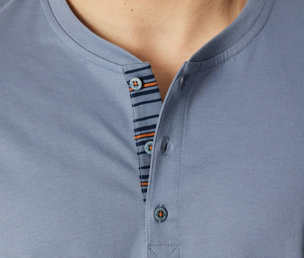 Nahaufnahme einer Person in einem blaugrauen SCHIESSER Shorty-Set Oberteil, das den Henley-Ausschnitt mit drei Knöpfen und eine gestreifte Knopfleiste in Dunkelblau und Orange zeigt.