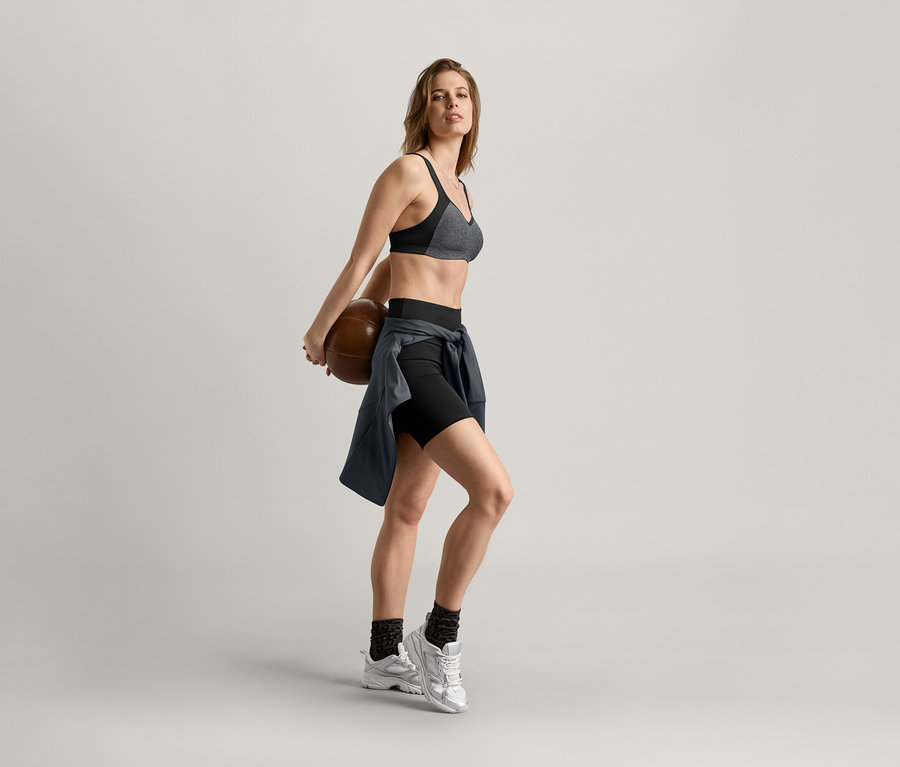 Frau hält einen Basketball hinter ihrem Rücken, trägt einen Sport-BH, Shorts und eine anthrazitfarbene Sportjacke.
