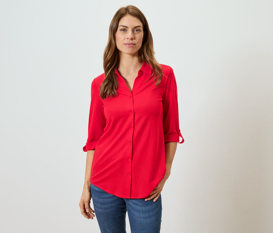 Frau mit braunen Haaren trägt eine rote Bluse und blaue Jeans.