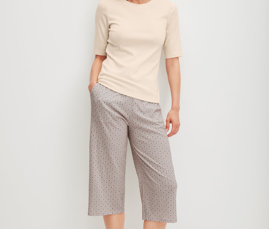 Frau steht barfuß, trägt ein helles T-Shirt und gemusterte Dreiviertelhosen.