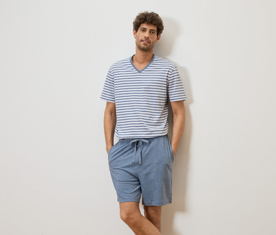 Ein Mann mit lockigem Haar, der ein gestreiftes T-Shirt und blaue Shorts trägt, steht vor einer weißen Wand.