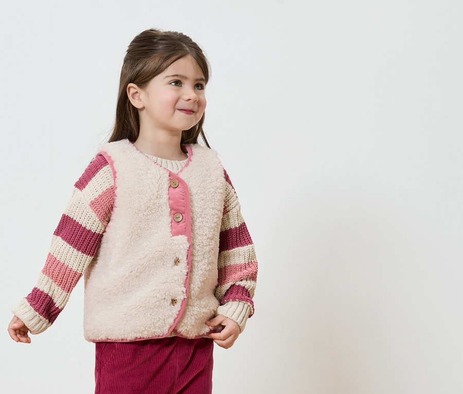 Lächelndes Mädchen mit gestreiftem Pullover und beiger Weste mit rosa Akzenten vor einem weißen Hintergrund.