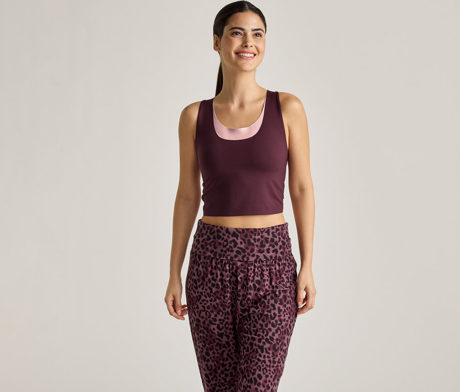 Frau mit Pferdeschwanz trägt bordeauxfarbenes Tanktop über einem rosa Top und einer Hose mit Leopardenmuster vor einem hellen Hintergrund.