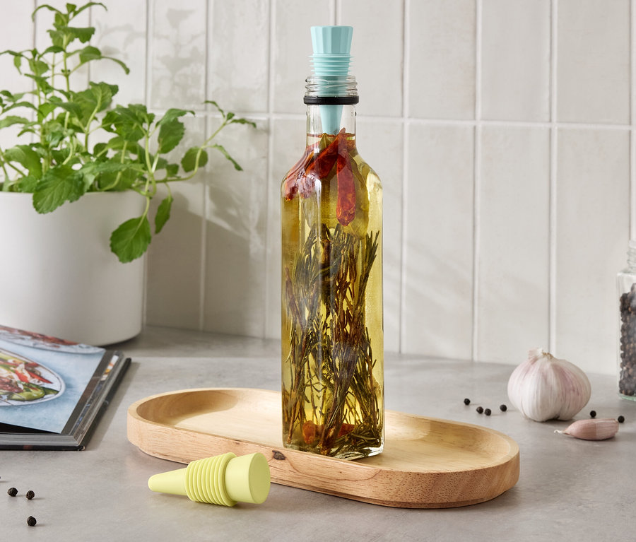 Eine Flasche mit Kräuteröl steht auf einem Holztablett, neben einer Knoblauchzehe und einem Glas mit Pfefferkörnern.