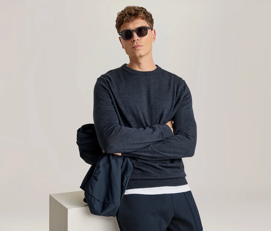 Mann mit lockigem braunem Haar und Sonnenbrille, der direkt in die Kamera blickt. Er trägt ein dunkelblaues Langarmshirt, ein weißes T-Shirt und eine dunkelblaue Hose. Seine Arme sind verschränkt, und eine dunkelblaue Jacke hängt über seinem linken Arm, der auf einem weißen Würfel ruht.