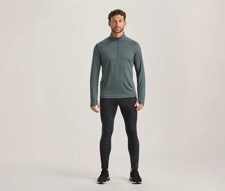 Mann steht in einem dunkelgrauen Langarm-Sportoberteil mit Reißverschluss und Daumenlöchern sowie schwarzen Sportleggings und schwarzen Sportschuhen.