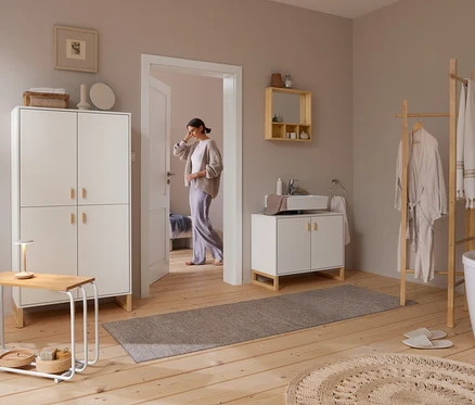 Eine Frau betritt ein helles Badezimmer mit Holzboden. Rechts steht eine Kleiderleiter mit Bademantel und Handtüchern neben einer weißen Badewanne. Über einem weißen Waschtisch mit Waschbecken hängt ein Spiegel aus massivem Bambus mit Ablagen. Links steht ein hoher weißer Schrank neben einem kleinen Tisch mit Lampe.
