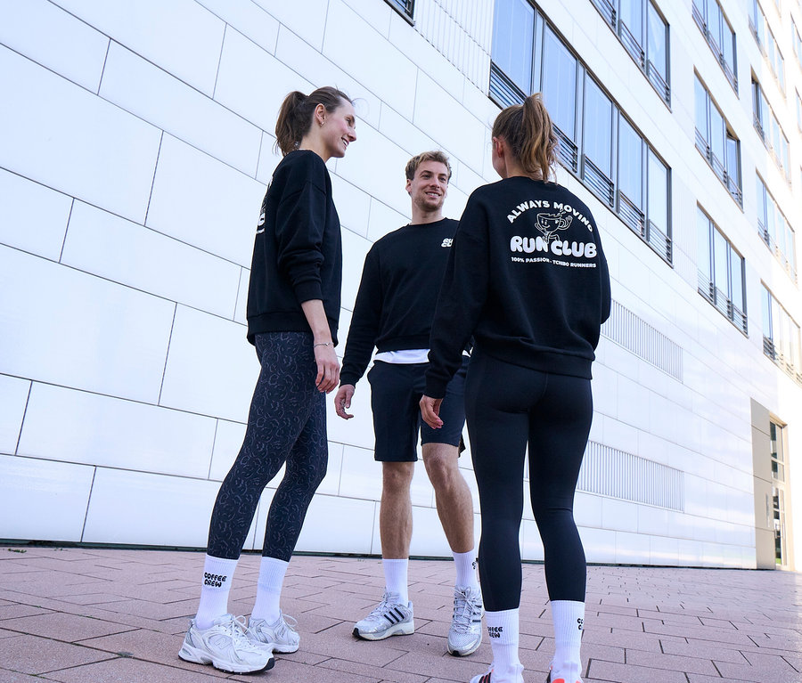 Drei Personen in schwarzen Sweatshirts und Sportkleidung unterhalten sich. Eine Frau trägt grau-schwarze Sportleggings »ActivePower« und Unisex Coffee Crew Tennissocken. Ein Mann trägt schwarze Sportshorts und weiße Socken. Eine andere Frau trägt schwarze Sportleggings »ActivePower« und ein Sweatshirt mit „RUN CLUB“-Logo.