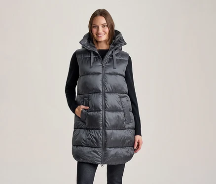 Lächelnde Frau trägt eine lange, dunkelgraue Kapuzen-Steppweste über einem schwarzen Langarmshirt und dunklen Hosen, eine Hand in einer Westentasche.
