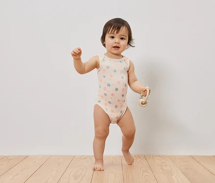 Ein Baby steht barfuß auf einem Holzboden, trägt einen hellrosa ärmellosen Baby-Body mit blauen und orangen Sternen und Muscheln und hält eine helle Rassel.