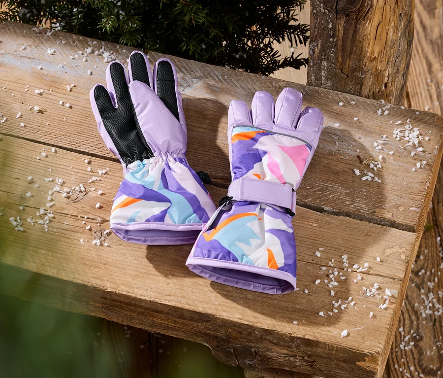 Zwei lilafarbene Handschuhe mit buntem, abstraktem Muster liegen auf einem Holztisch mit Kunstschnee und Tannenzweigen.