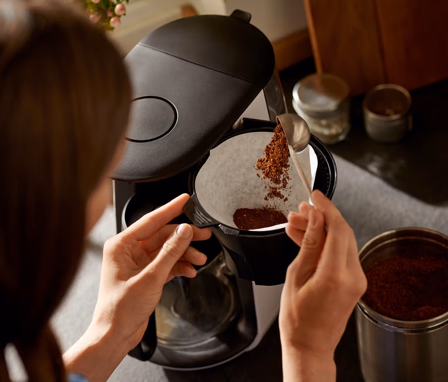 Frau füllt mit einem Löffel Kaffee in den weißen Filter der »Let's Brew« Filterkaffeemaschine.