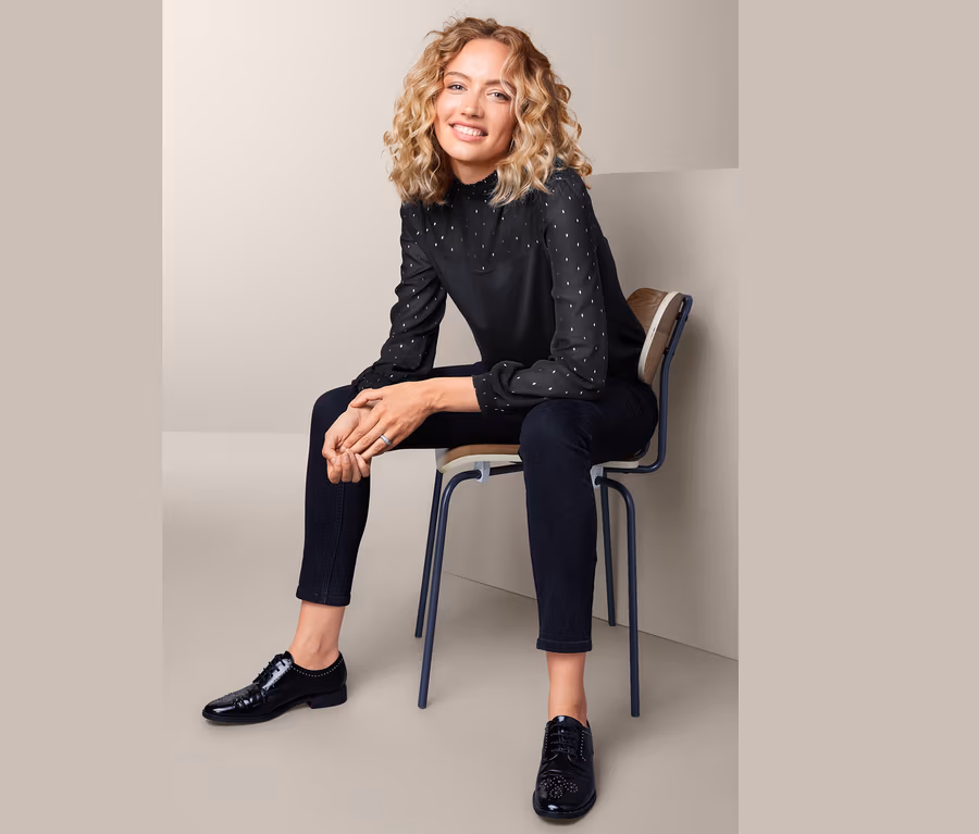 Frau sitzt auf einem Stuhl und trägt ein Blusenshirt im Materialmix, schwarze Hose und schwarze Schuhe.