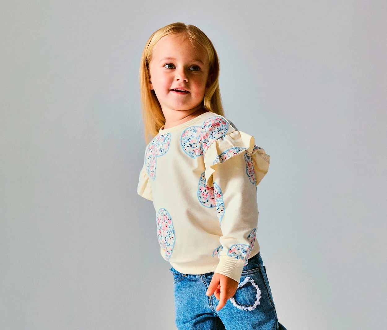 Junges Mädchen mit blondem Haar trägt einen cremefarbenen NAME IT Kinder-Sweater mit floralen Herzmustern und Rüschen an den Schultern, dazu blaue Jeans, blickt nach rechts.