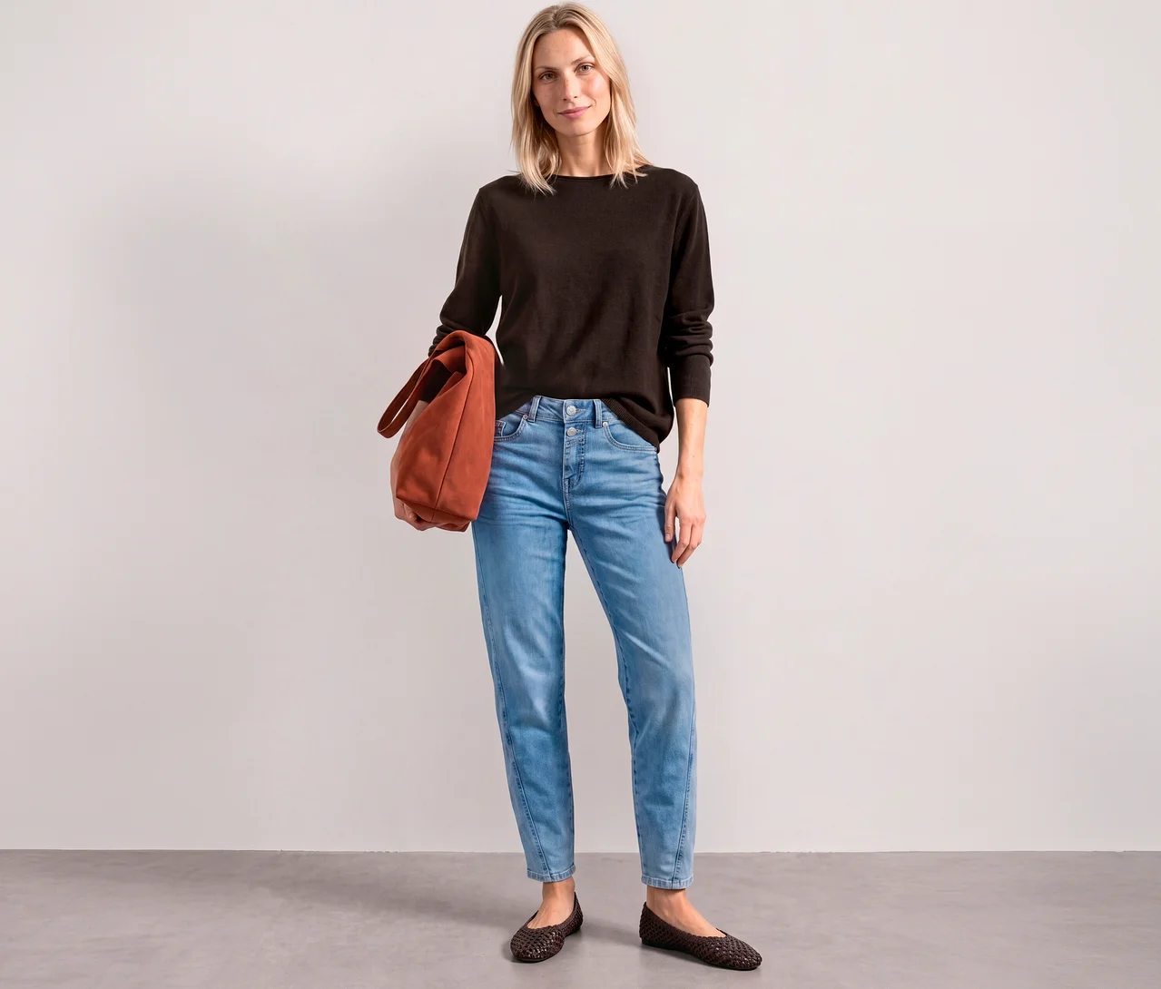 Eine Frau trägt einen braunen Street One Strickpullover, hellblaue Jeans und hält eine orangefarbene Tasche in der Hand.