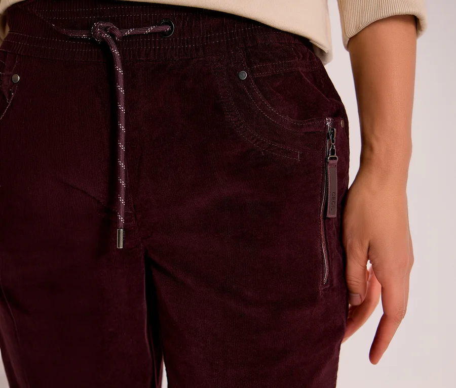Nahaufnahme einer Person in einer bordeauxroten CECIL Cordhose mit elastischem Bund, gesprenkeltem Kordelzug und Reißverschlusstasche mit CECIL Etikett.