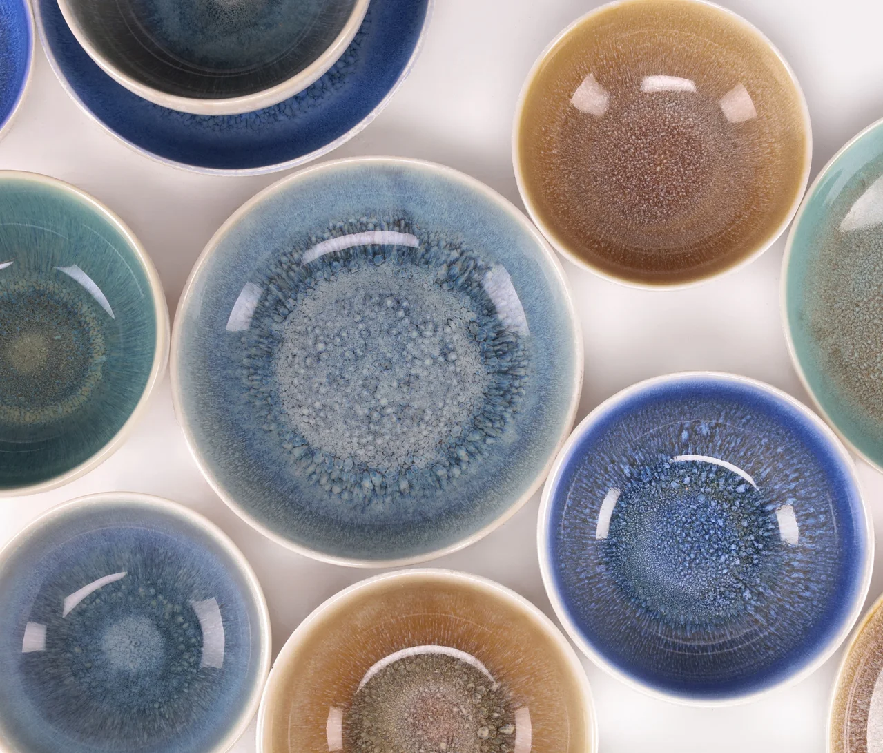 Eine Draufsicht auf mehrere Keramikschalen aus dem creaTable Bowl-Set "Caldera", mit einer glänzenden, gesprenkelten Glasur in verschiedenen Blau-, Türkis- und Braungoldtönen.
