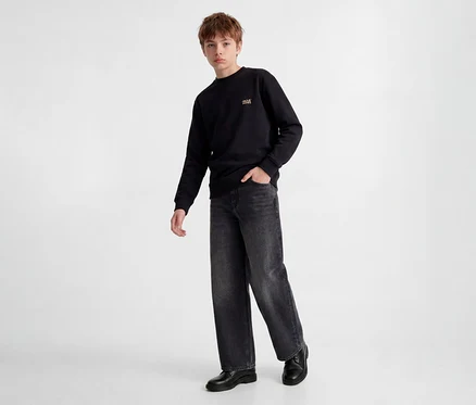Junger Junge steht in schwarzem Sweatshirt mit kleinem goldenem Logo, dunkelgrauen Wide-Leg Jeans und schwarzen Schuhen. Eine Hand steckt in der Hosentasche.