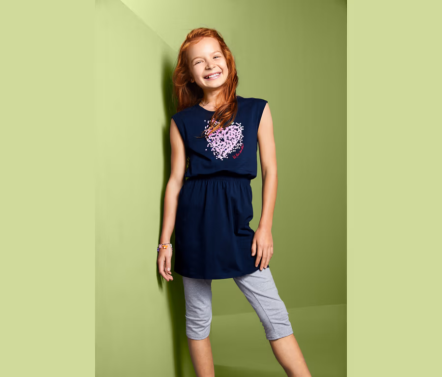 Lächelndes Mädchen posiert in marineblauem Jerseykleid mit Wendepailletten und grauen Leggings.