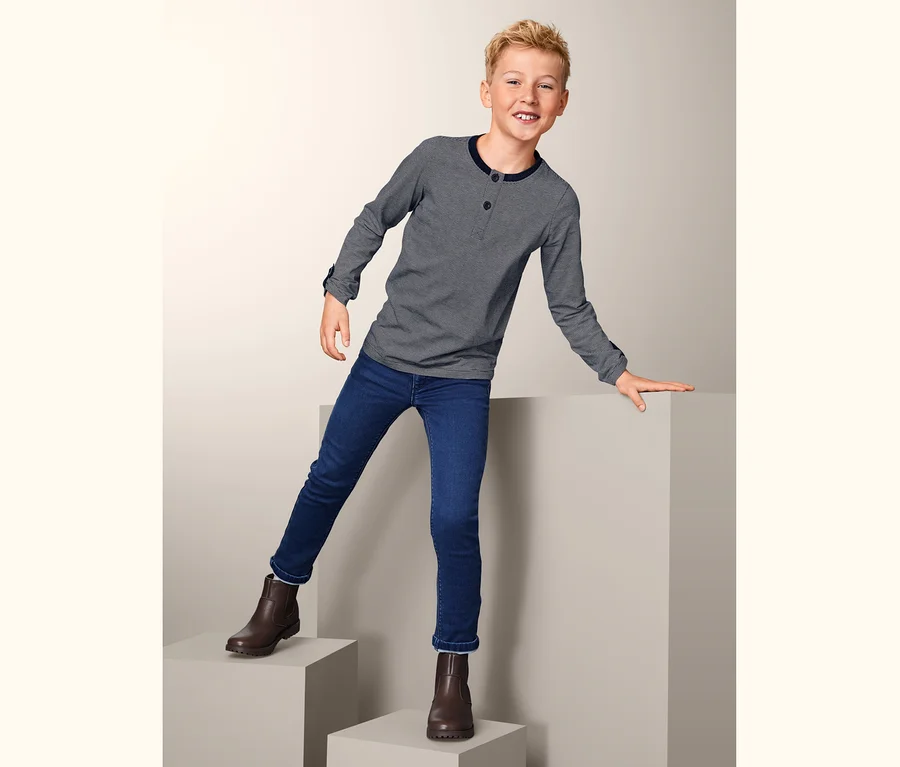 Junge steht auf grauen Würfeln und trägt Jeans und 2 Langarmshirts.