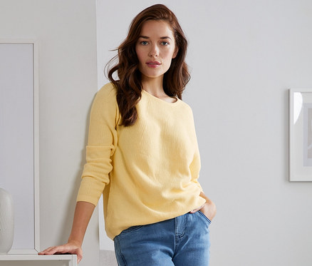 Eine Frau lehnt an einem weißen Bücherregal und trägt einen gelben Strickpullover.
